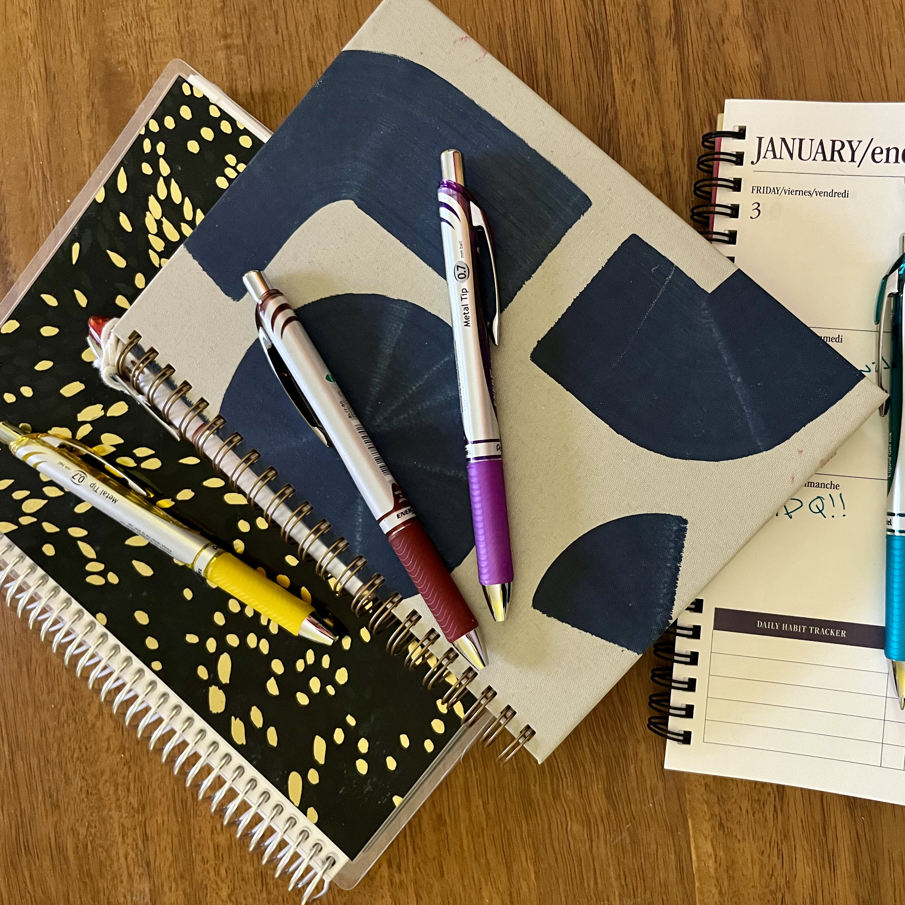 Several notebooks and pens in different colors sprawled across a wooden desk